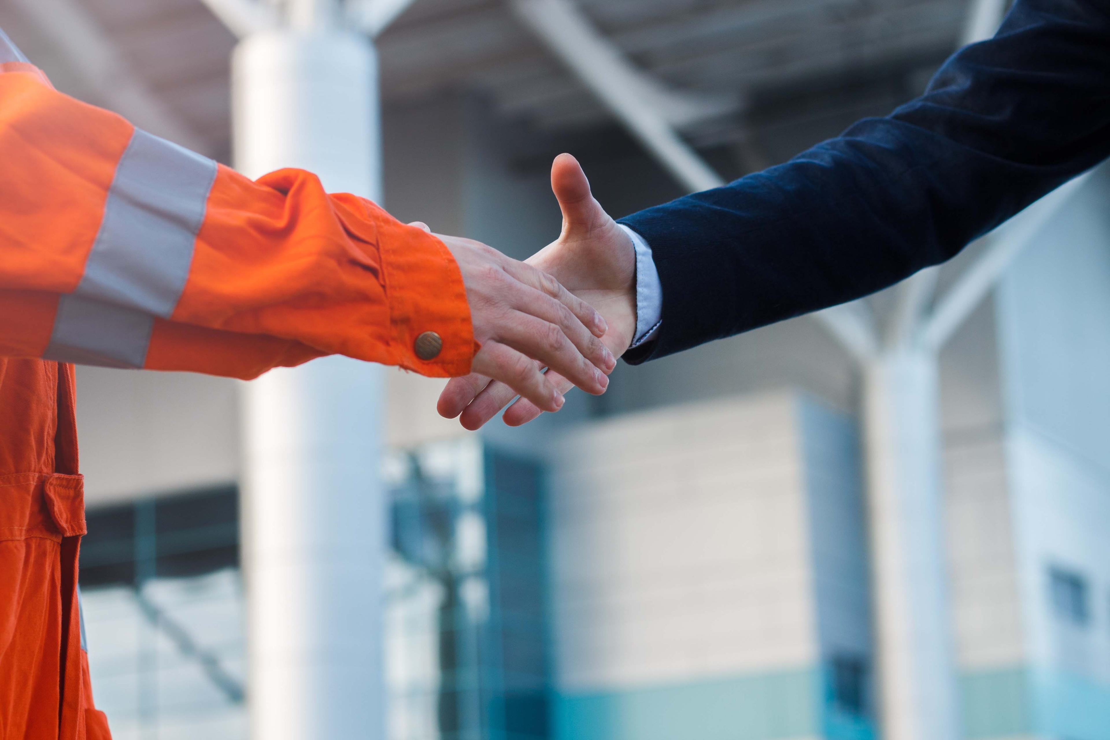 A mine worker wearing an orange high visibility shirt and an office worker wearing business attire shaking hands.