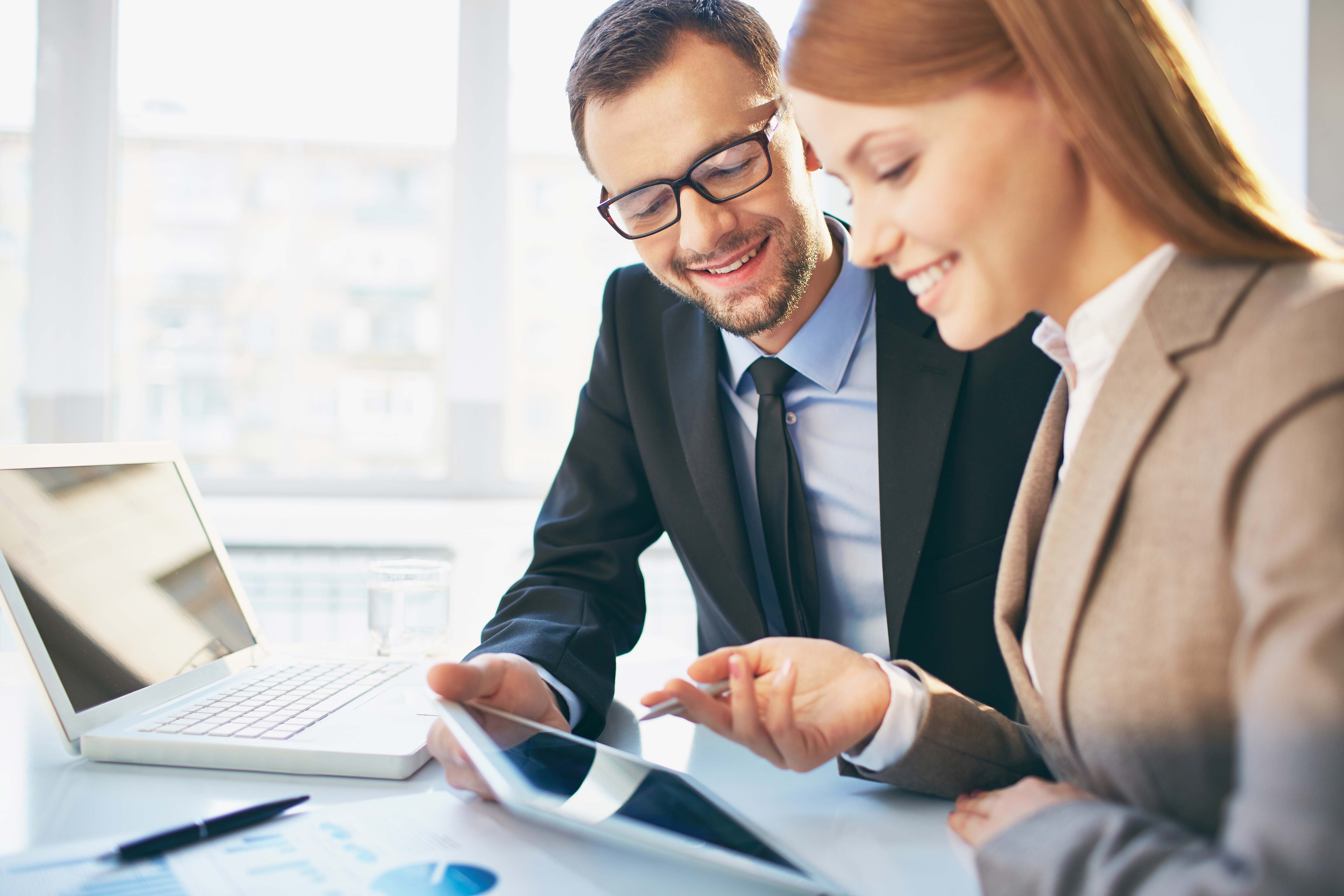 Smiling office workers wearing business attire looking at a laptop and mobile device.