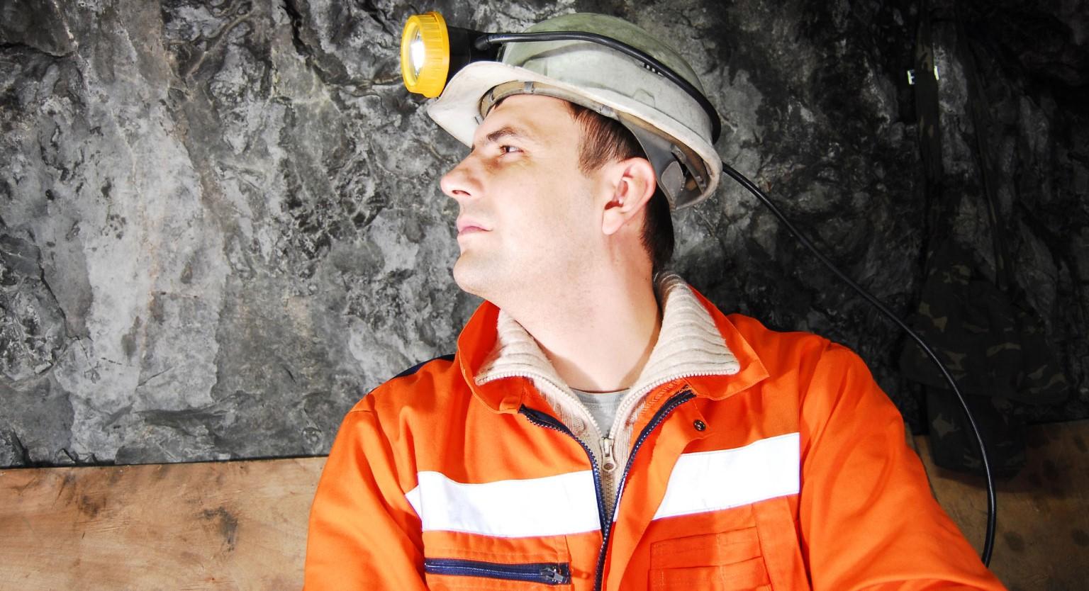 Underground mine worker wearing an orange high visibility shirt and a white hard hat with a headlamp.